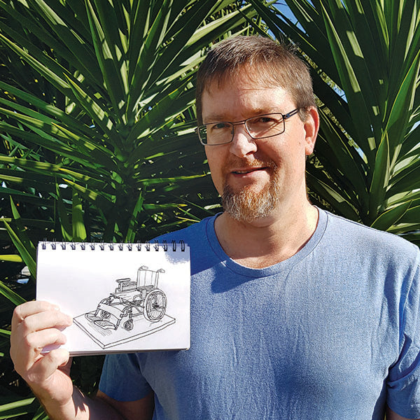 A man in a blue shirt holds a sketchpad with a black and white drawing of a wheelchair-like structure. He stands in front of tall green plants, smiling at the camera.