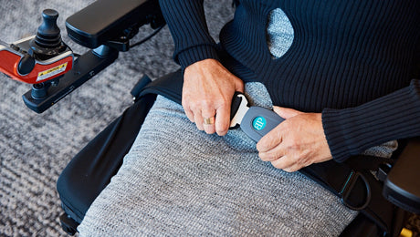 A person in a wheelchair fastens a seatbelt across their lap, wearing a grey skirt and black top. One hand holds the buckle while the other guides the belt.