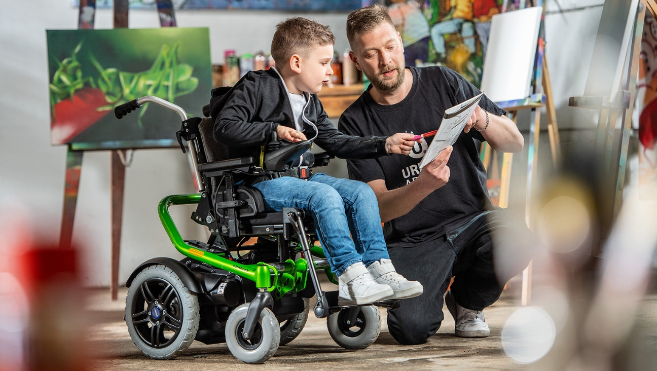 A young boy in a wheelchair and an adult man kneel together in an art studio, looking at a drawing. Colorful paintings and art supplies are visible in the background.
