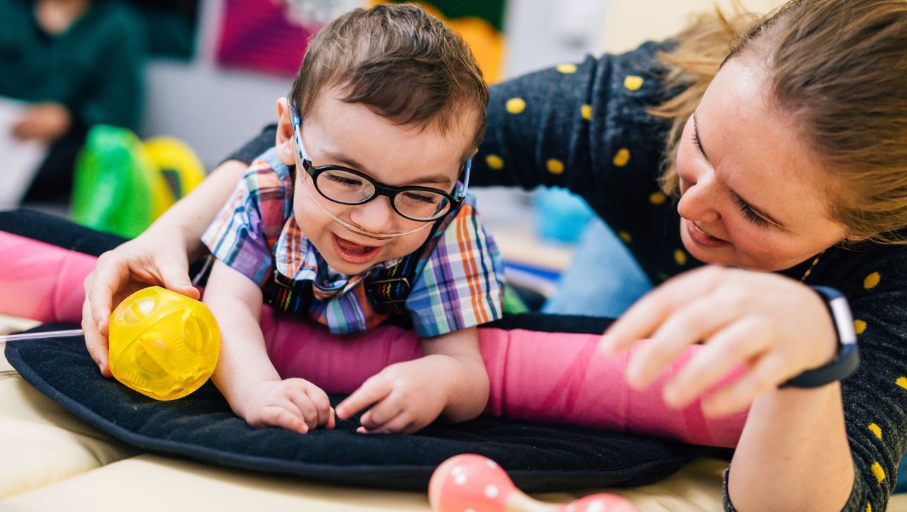 A young boy wearing glasses and a plaid shirt smiles while playing with a yellow ball, supported by a woman who is smiling and encouraging him. They are in a colorful, playful environment.