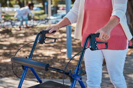 A person in a pink top and white pants uses a blue walker outdoors, grasping the handles. They are standing on a wooden path with trees and another person blurred in the background.