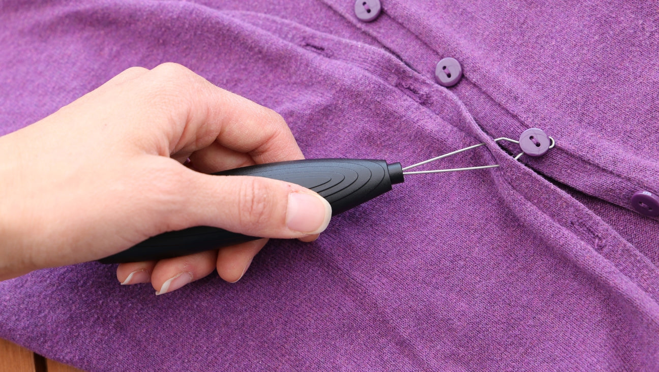 A hand uses a black sewing tool to thread a purple button onto a matching purple garment with several buttons already attached.