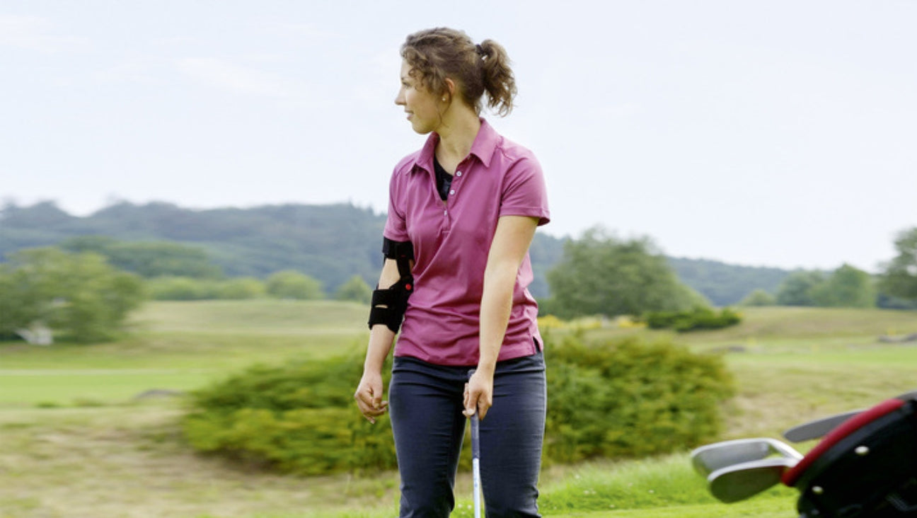 A woman wearing a pink polo shirt and an arm brace stands on a golf course, holding a golf club and looking to the side. Golf equipment and green landscape are visible.