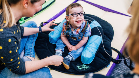 A smiling child with glasses is seated on a supportive cushion. The child is wearing a plaid shirt and overalls. A caregiver interacts with the child, who is surrounded by soft, colorful equipment.