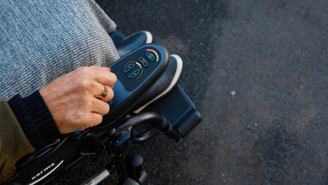 A close-up of a persons hand operating the control panel of an electric wheelchair outdoors. The person has a blanket over their lap.