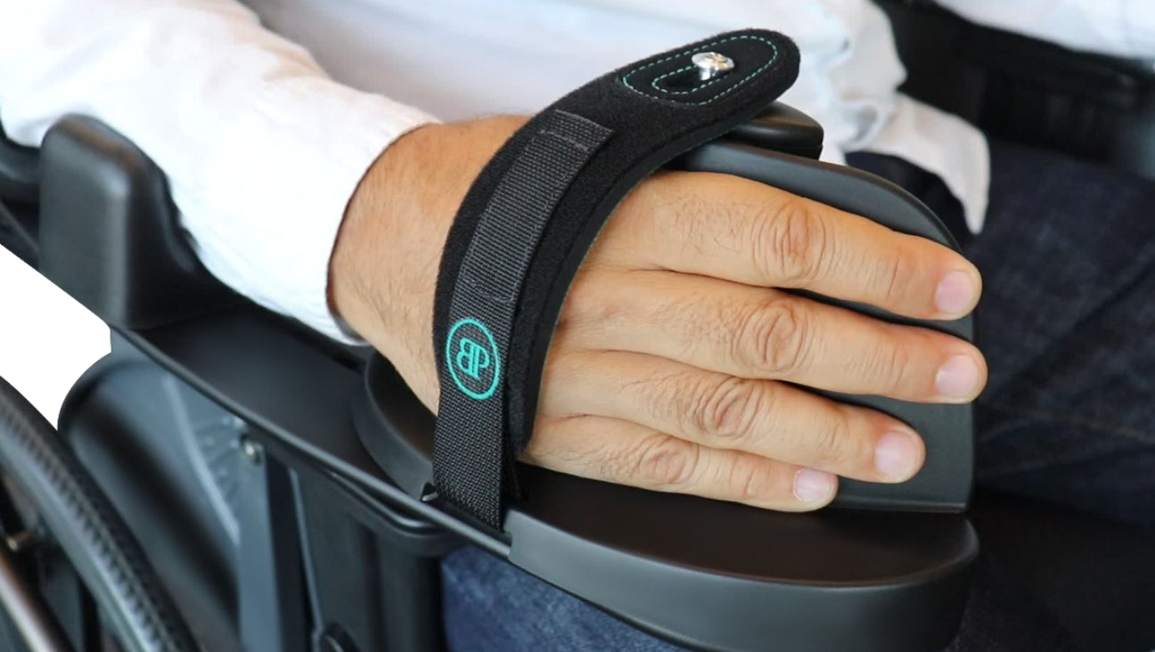 A person’s hand secured to a wheelchair armrest with a black strap, providing support and stability. The strap features a blue embroidered logo.