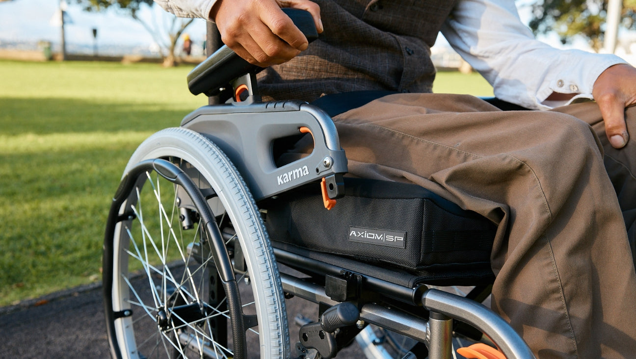 A person in brown pants sits in a wheelchair on a sunny day in a park, with one hand resting on the armrest and green grass in the background.
