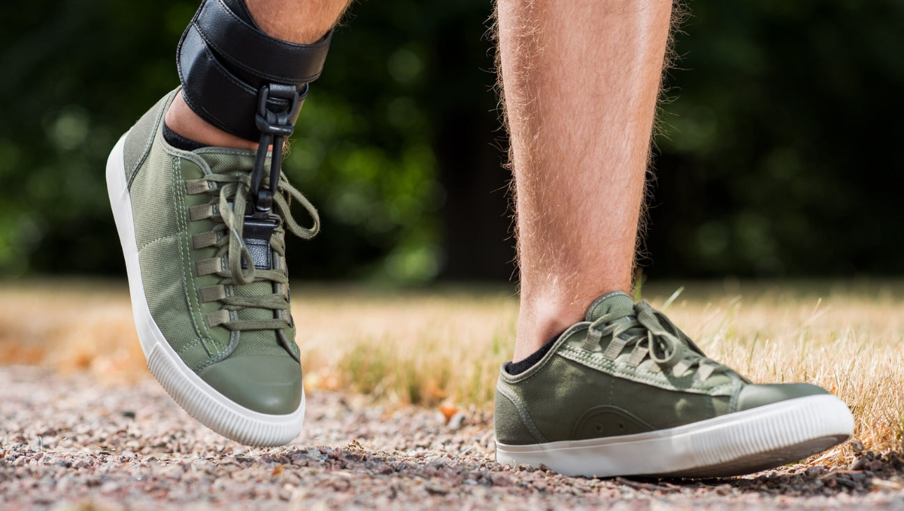 A person walks outdoors on a gravel path wearing green sneakers and an ankle-foot orthosis on one leg, with grass and trees in the blurred background.