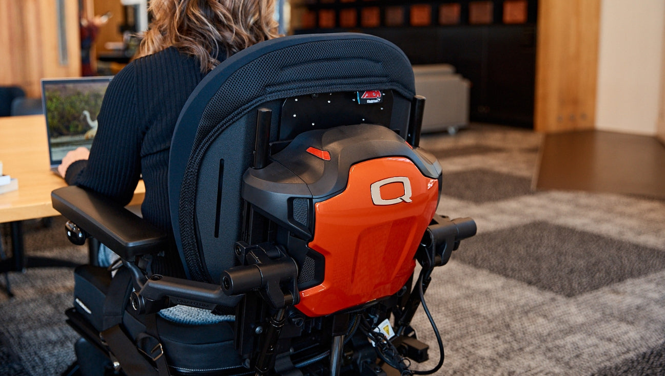 Person sitting in a motorized wheelchair featuring a sleek, orange backing with a bold Q logo. They are positioned at a wooden desk, using a laptop in a modern office environment.