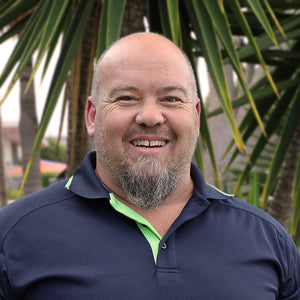 Man smiling outdoors, standing in front of a large palm-like plant. He is wearing a navy blue polo shirt with a light green collar. The background features blurred greenery and some structures.