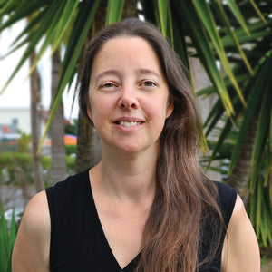 A person with long brown hair and a black sleeveless top stands outside, smiling. In the background, there are green palm leaves and a blurred view of buildings and a street.